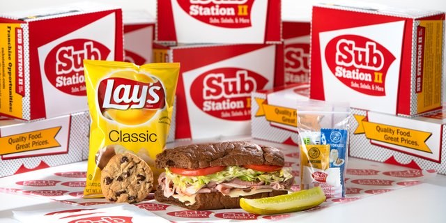 A Sub Station II box meal with a sub, Lay's chips, a cookie, a pickle and condiments, in front of stacked catering boxes